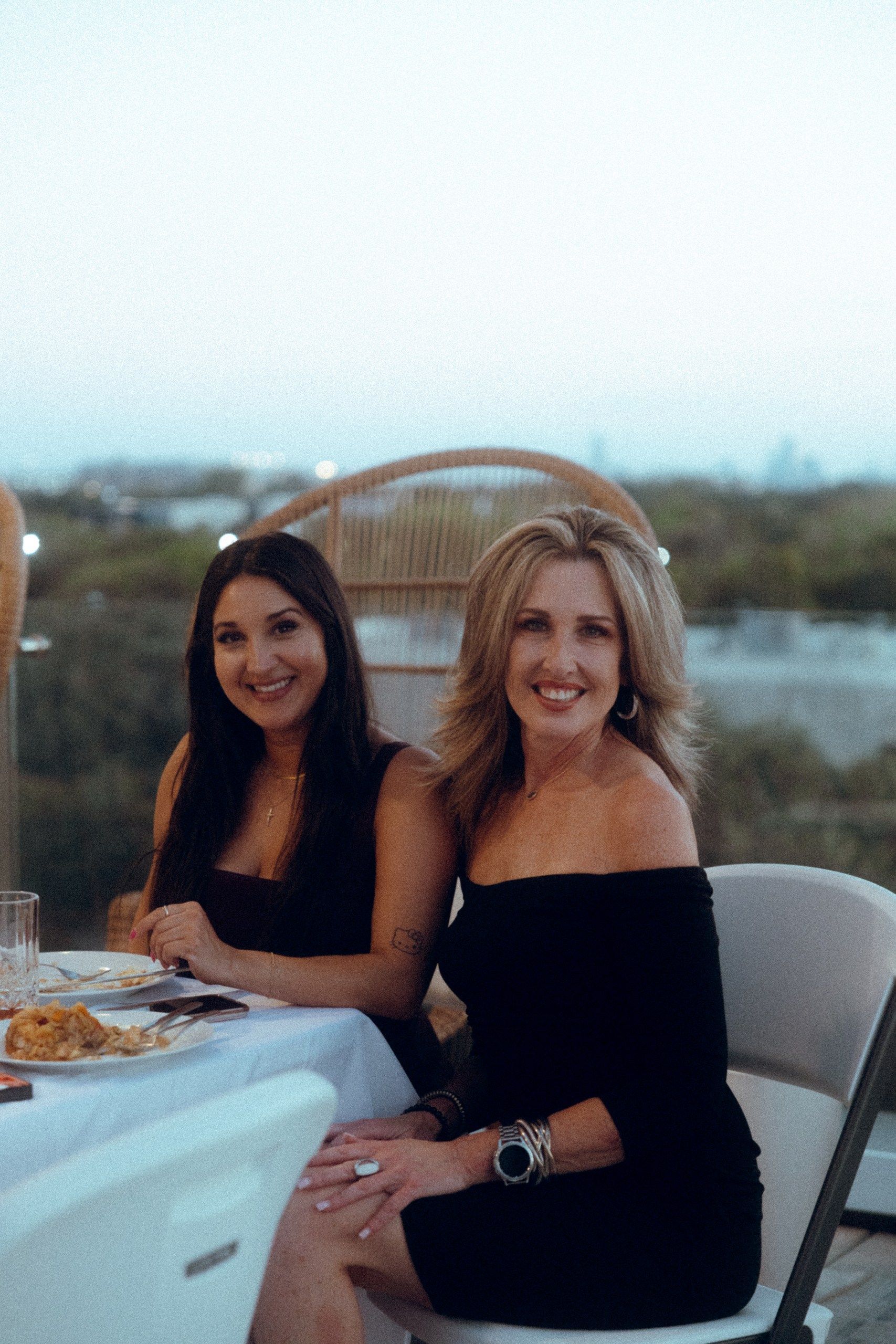 Two smiling women at an outdoor table. One wears an off-the-shoulder black dress, the other a black dress. Evening setting.