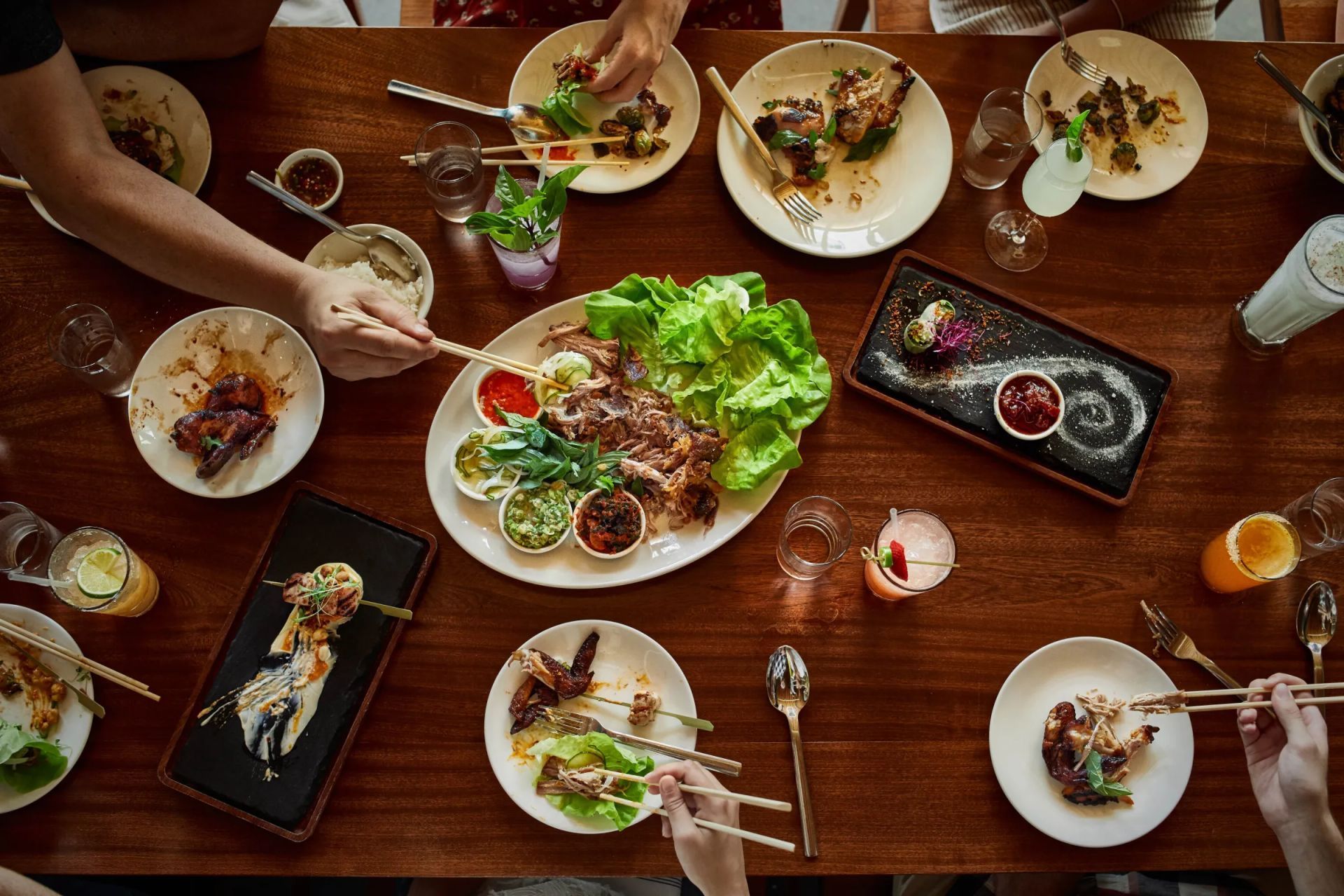 People eating Asian food at a wooden table with plates of food, lettuce wraps, and drinks.