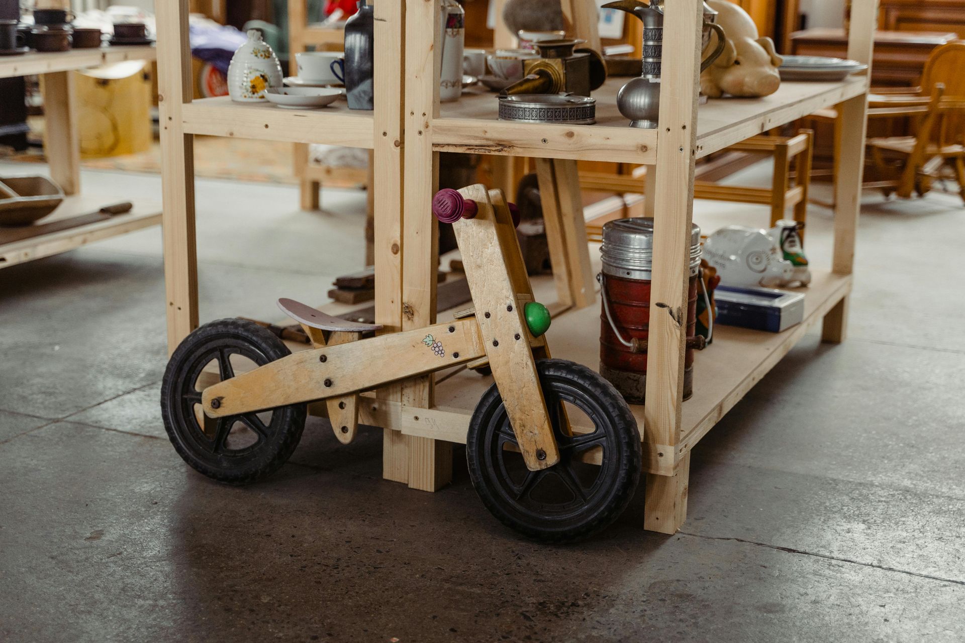 Moto giocattolo in legno, legno chiaro, ruote nere, posizionata sotto uno scaffale in legno su cui sono esposti vari oggetti.