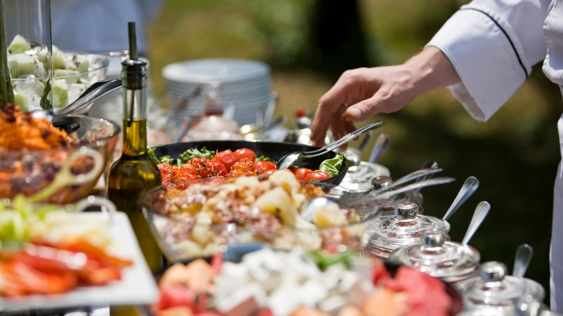 Buffet table with assorted dishes and a hand reaching to serve food outdoors