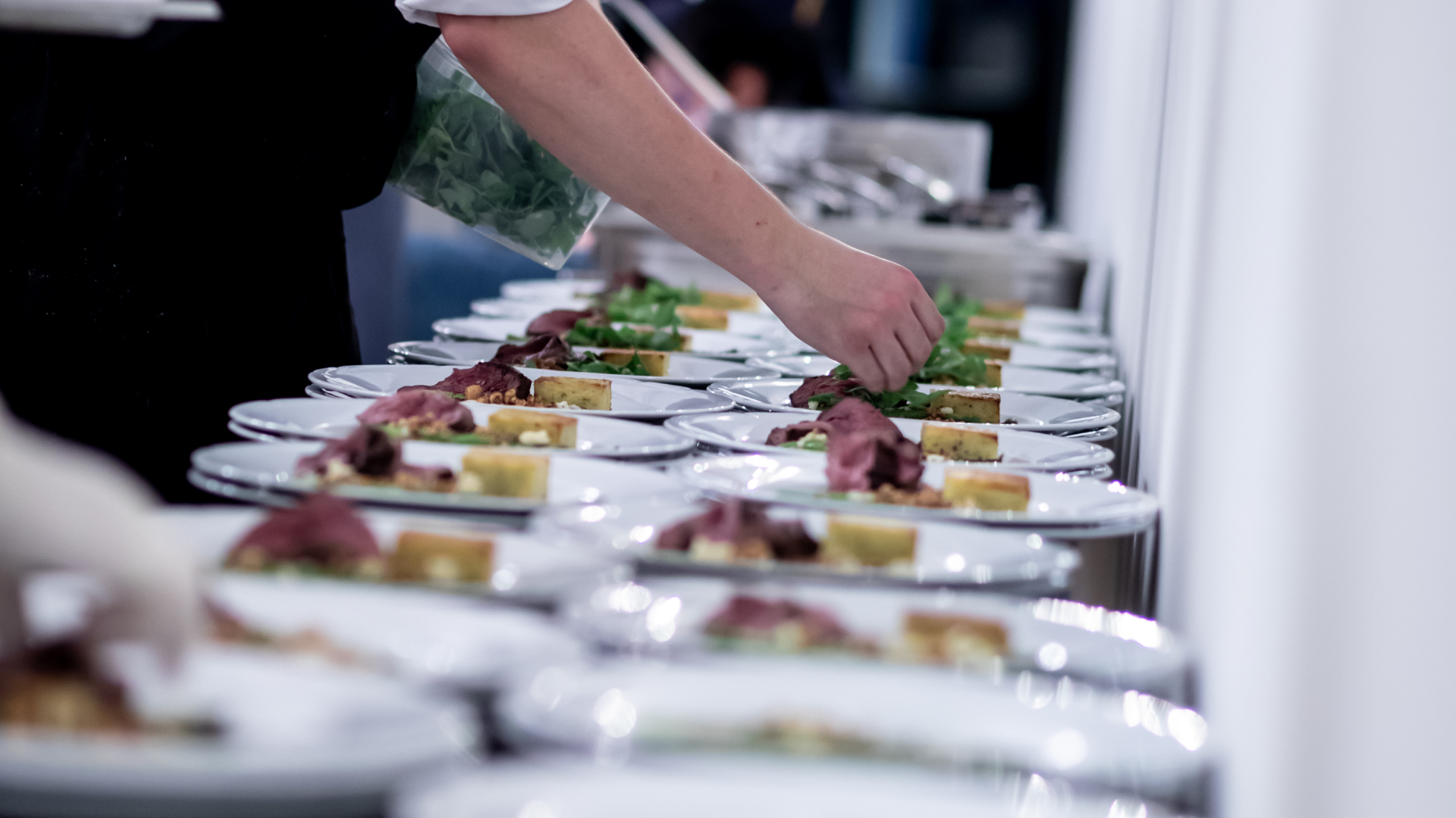 Hands arranging appetizers on a long buffet table with white plates and silver serving dishes