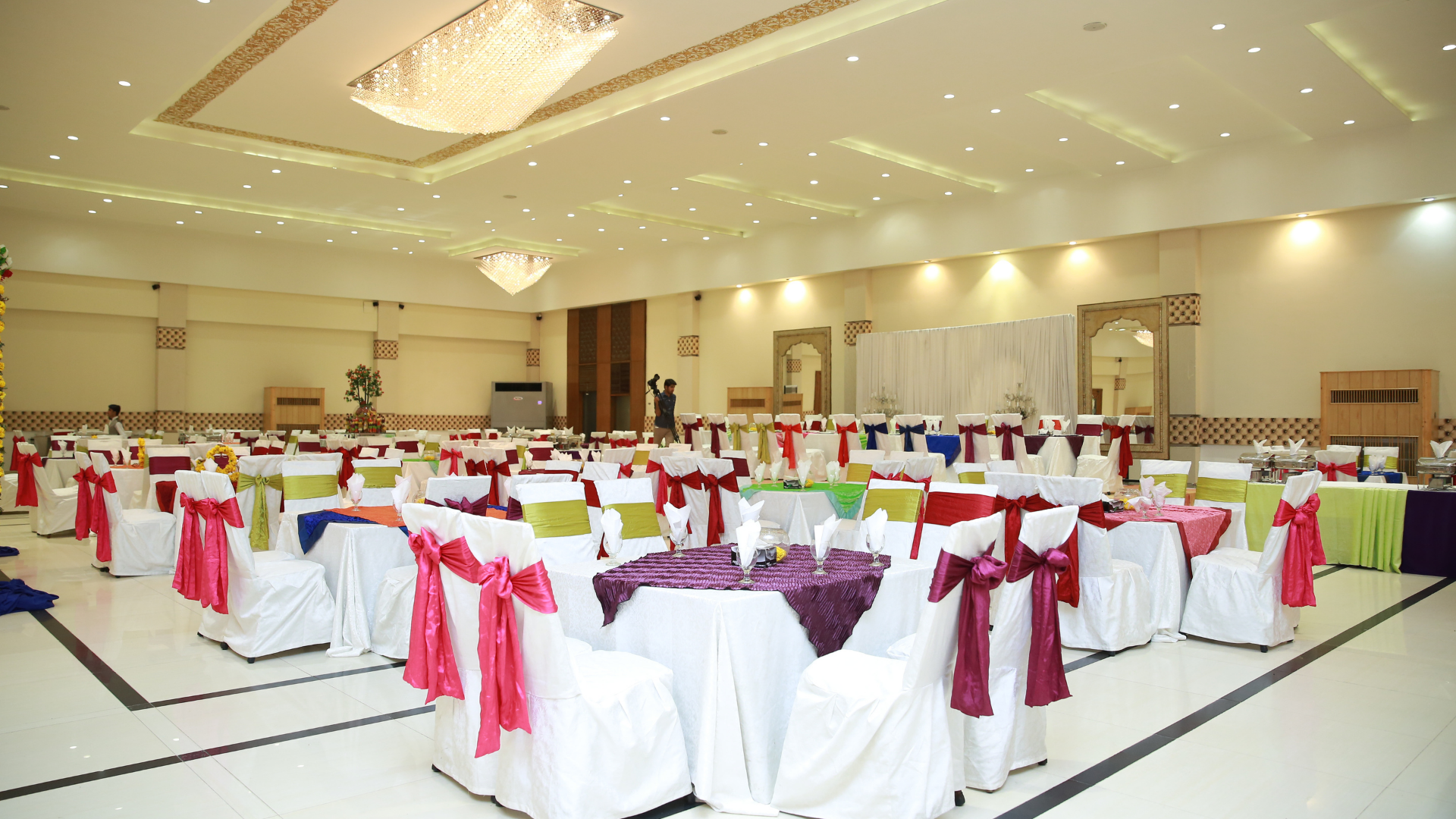 Banquet hall with rows of round tables dressed in white and burgundy chair covers under warm ceiling lights