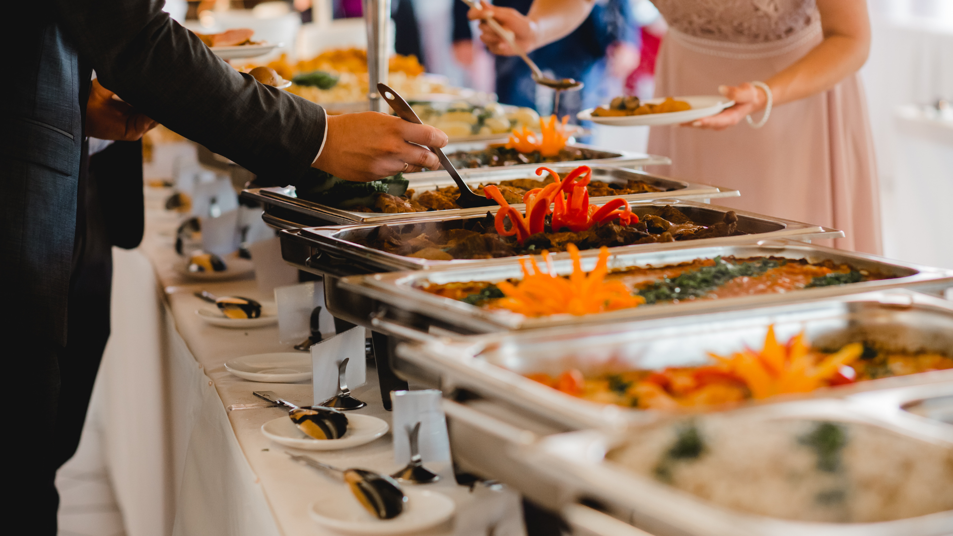 Guests serving buffet dishes at a catered event with trays of food and tongs