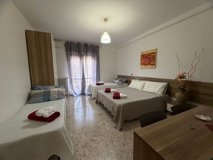 Piccola camera da letto con letto in legno, copriletto a fantasia, comodino e finestra con tende trasparenti.