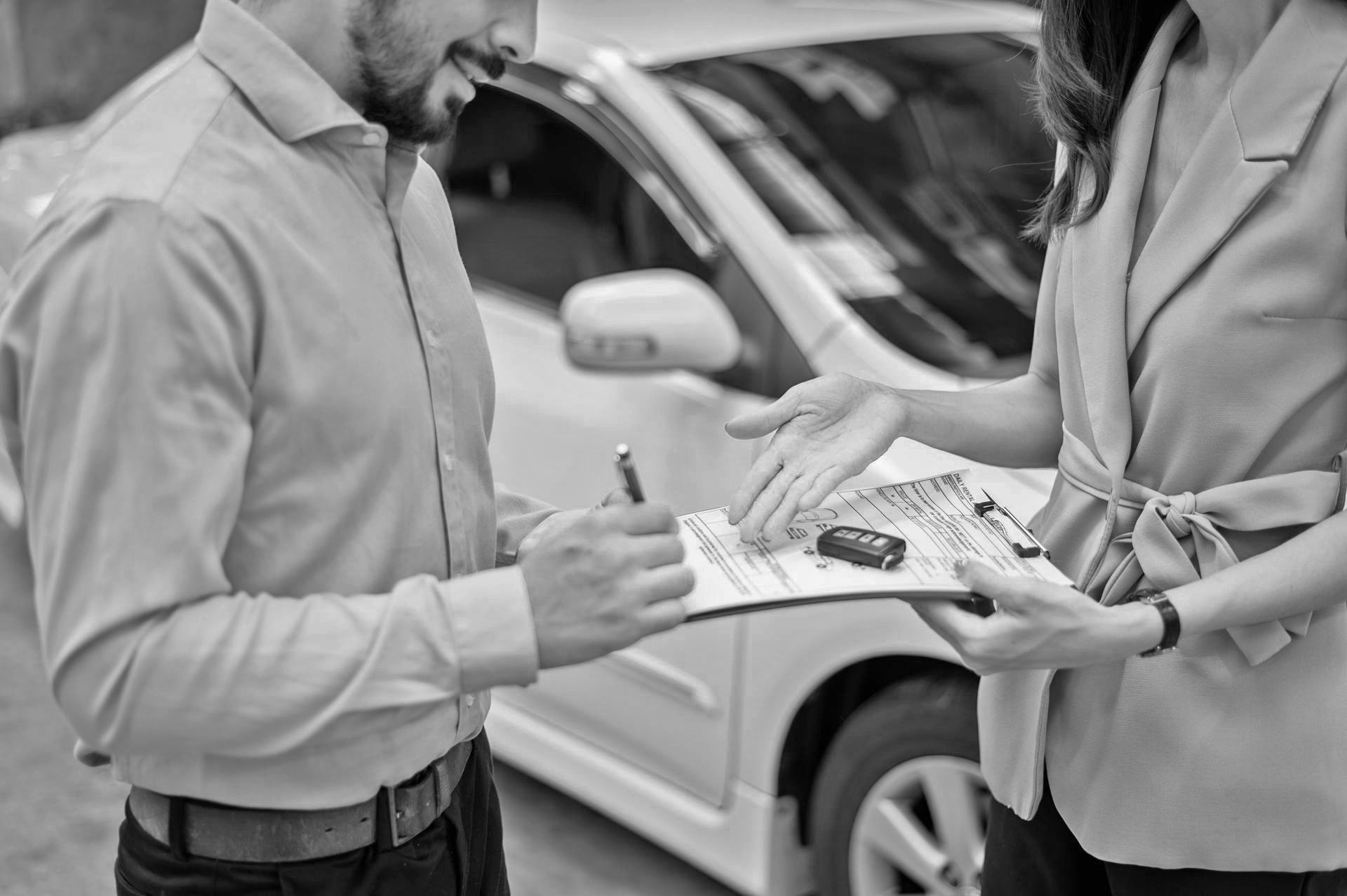 A man is signing a document while a woman holds a clipboard in front of a car.