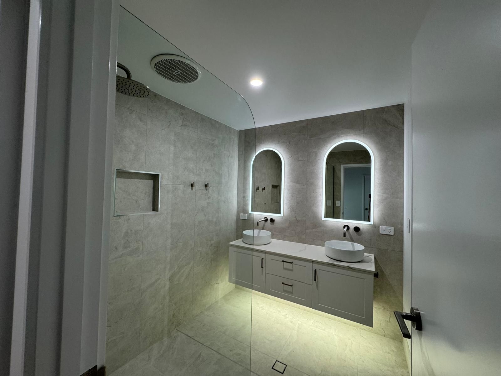 Modern bathroom with gray tiles, dual sinks, arched mirrors with lights, and a glass shower.