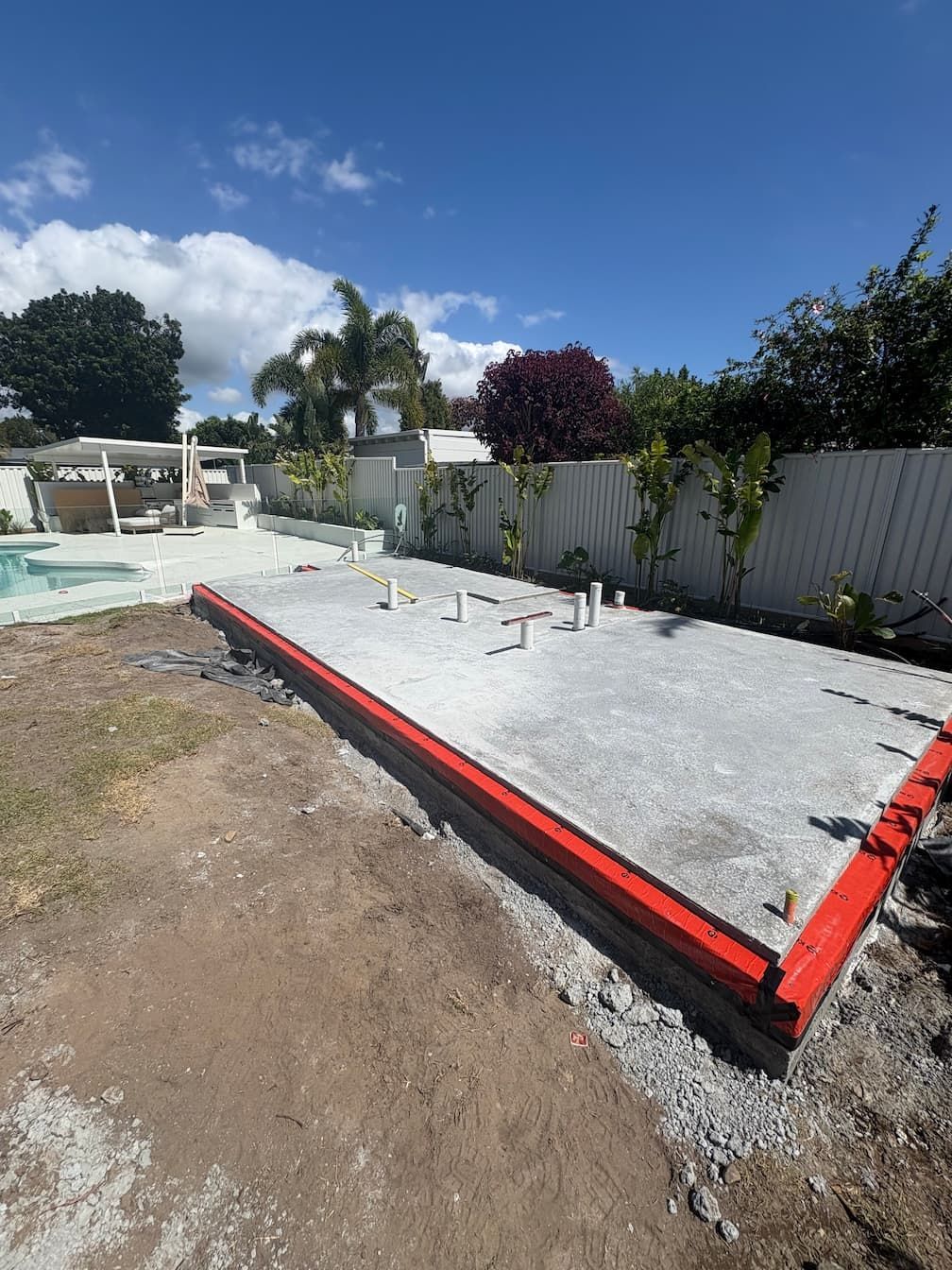 Construction site in a backyard. Concrete foundation with red border and pipes, gravel, and surrounding dirt.