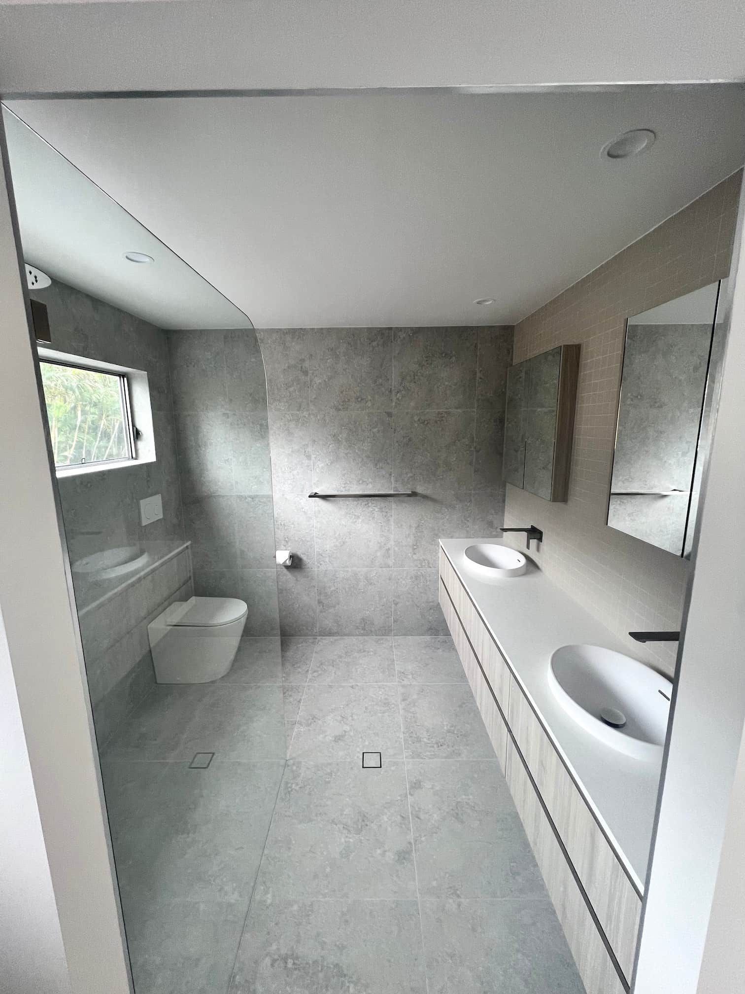 Modern bathroom with gray tiled walls and floor, toilet, double vanity, mirrors, and shower area.