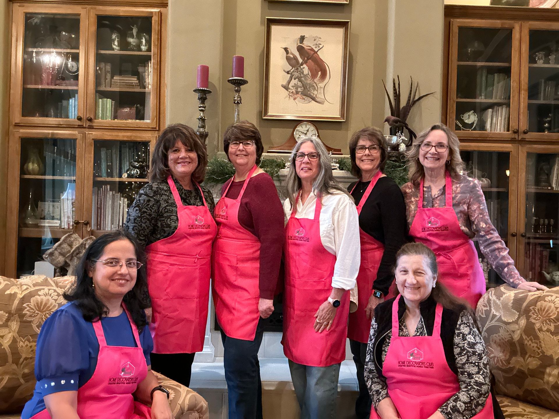 Group of women in pink aprons pose in a living room, smiling, some leaning.