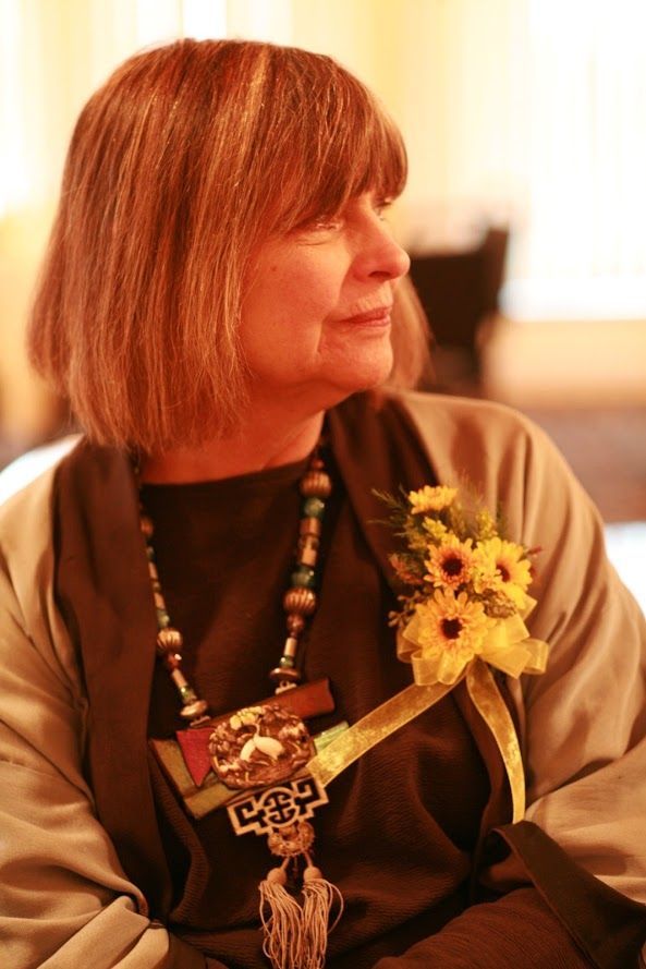 Woman with short brown hair, wearing a decorative necklace and flowers on her jacket.