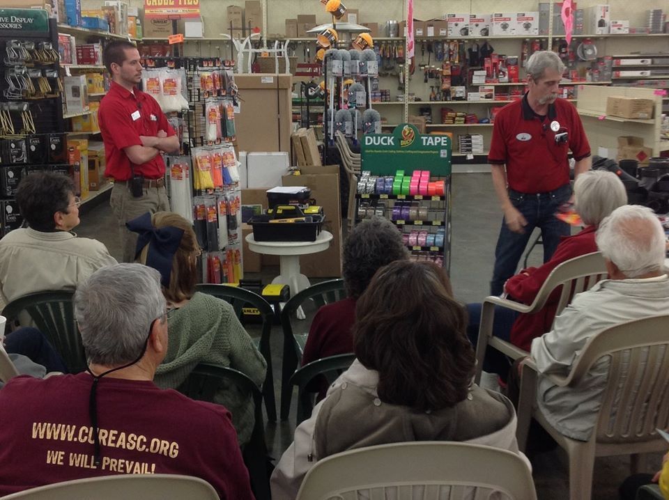 Two men in red shirts give a presentation to a seated group in a hardware store.