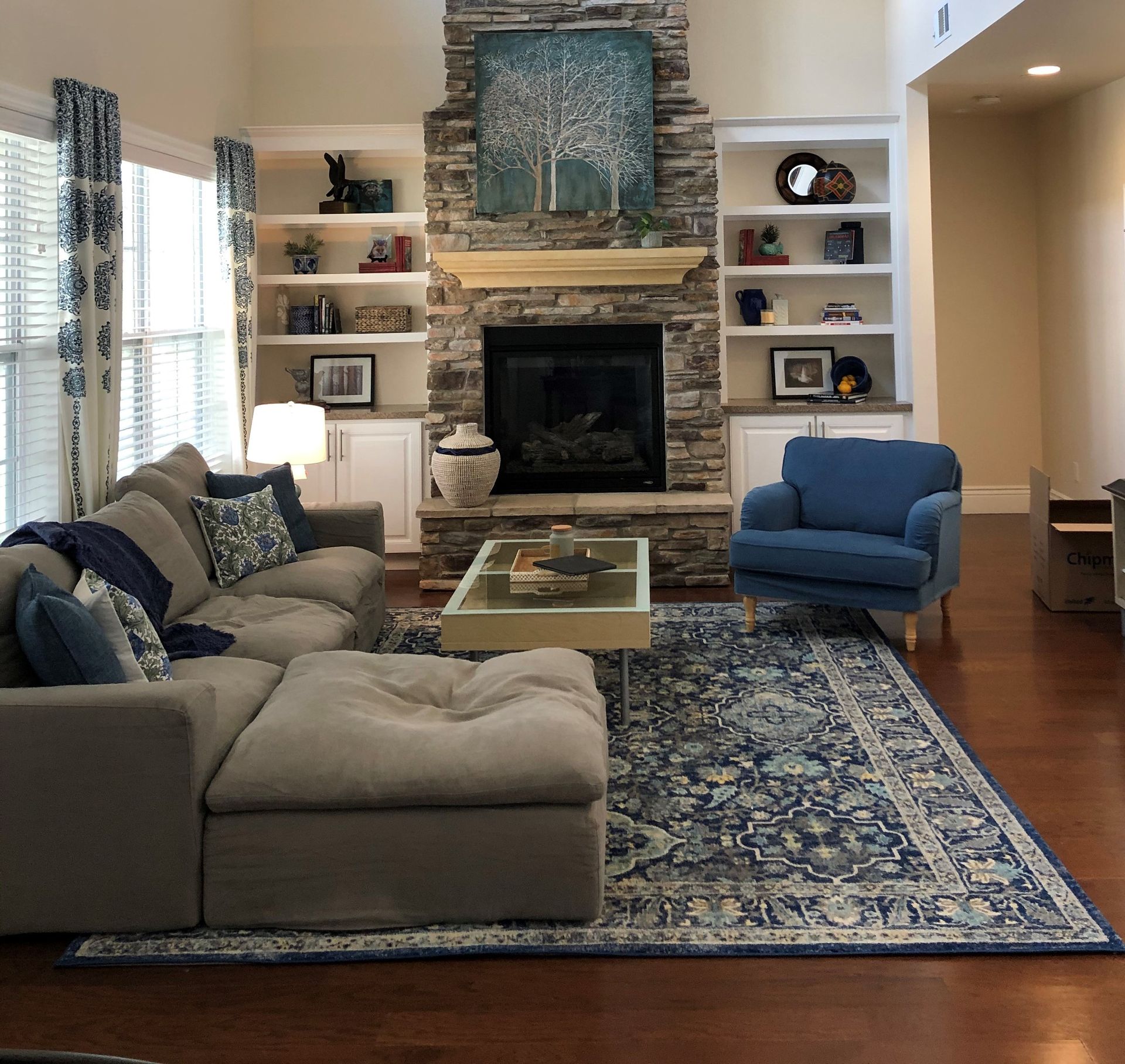 Living room with fireplace, built-in shelves, gray sectional, blue rug, armchair, and patterned curtains.