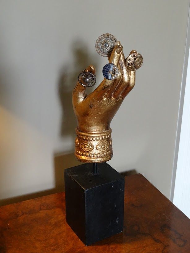 Golden hand sculpture holding rings on a black square base, on a wooden surface.