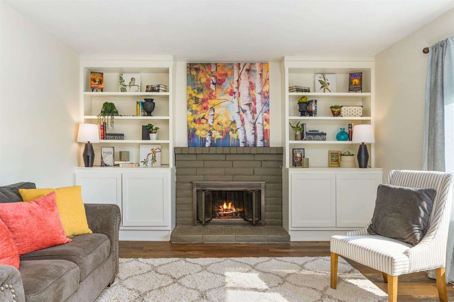 Living room with fireplace, bookshelves, and colorful artwork. A couch and patterned chair face the fireplace.