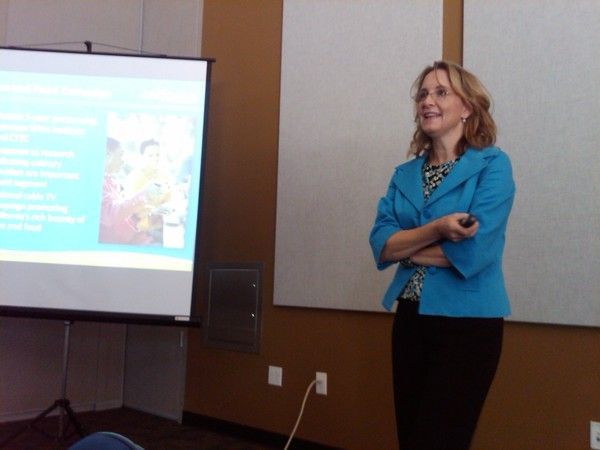 Woman in blue blazer presents in a classroom. A projector screen shows a slide with text and an image.