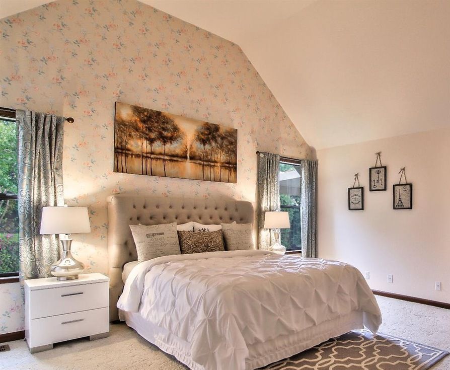Bedroom with a tufted headboard, floral wallpaper, artwork, and white bedding; soft lighting.
