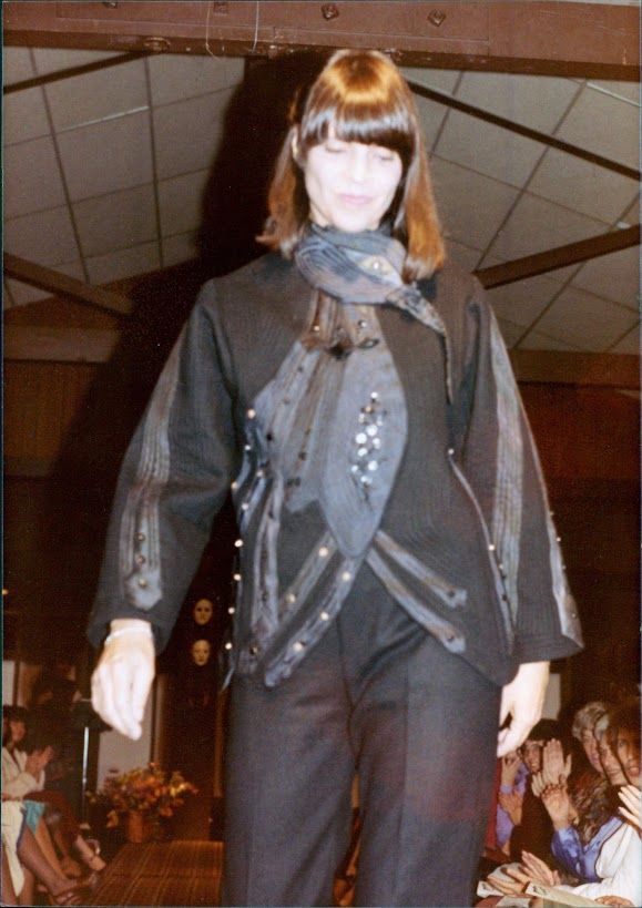 Woman in black outfit, walking on a runway; brown bangs, black jacket with accents, black pants.