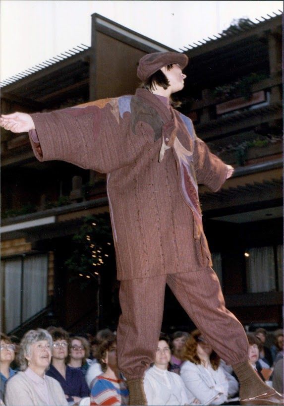 Model in brown jumpsuit and cap poses with arms outstretched on a platform, in front of an audience.