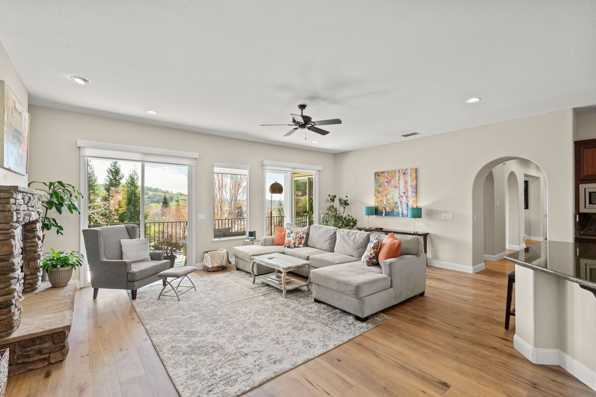 Living room with gray sectional sofa, fireplace, hardwood floors, and windows overlooking trees.