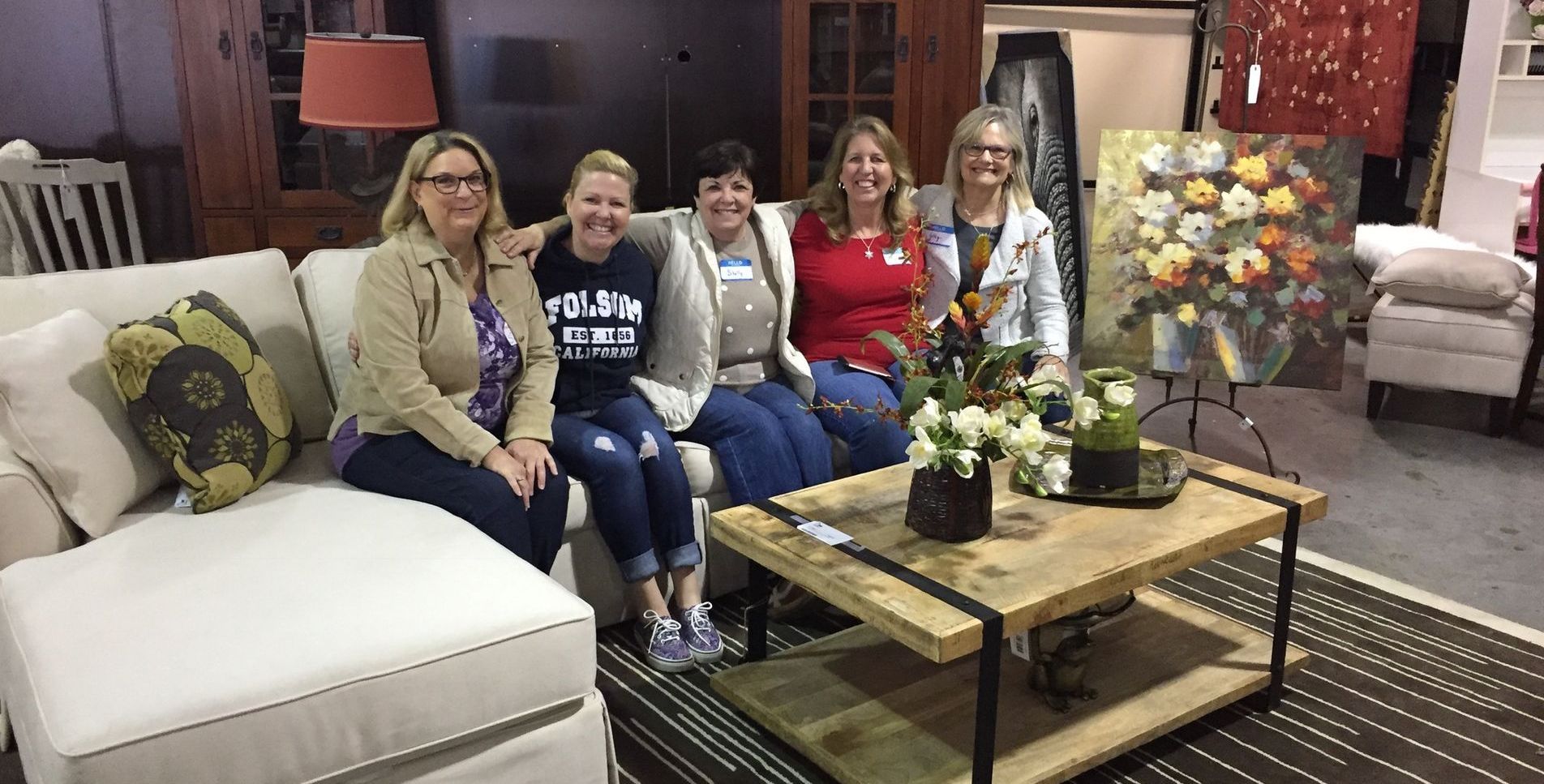 Five women sit on a cream sofa in a furniture store. A wood and metal coffee table sits in front of them.