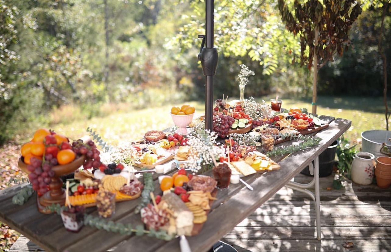 Picnic table laden with food in a sunny outdoor setting. Fruits, cheese, and treats arranged for a gathering.