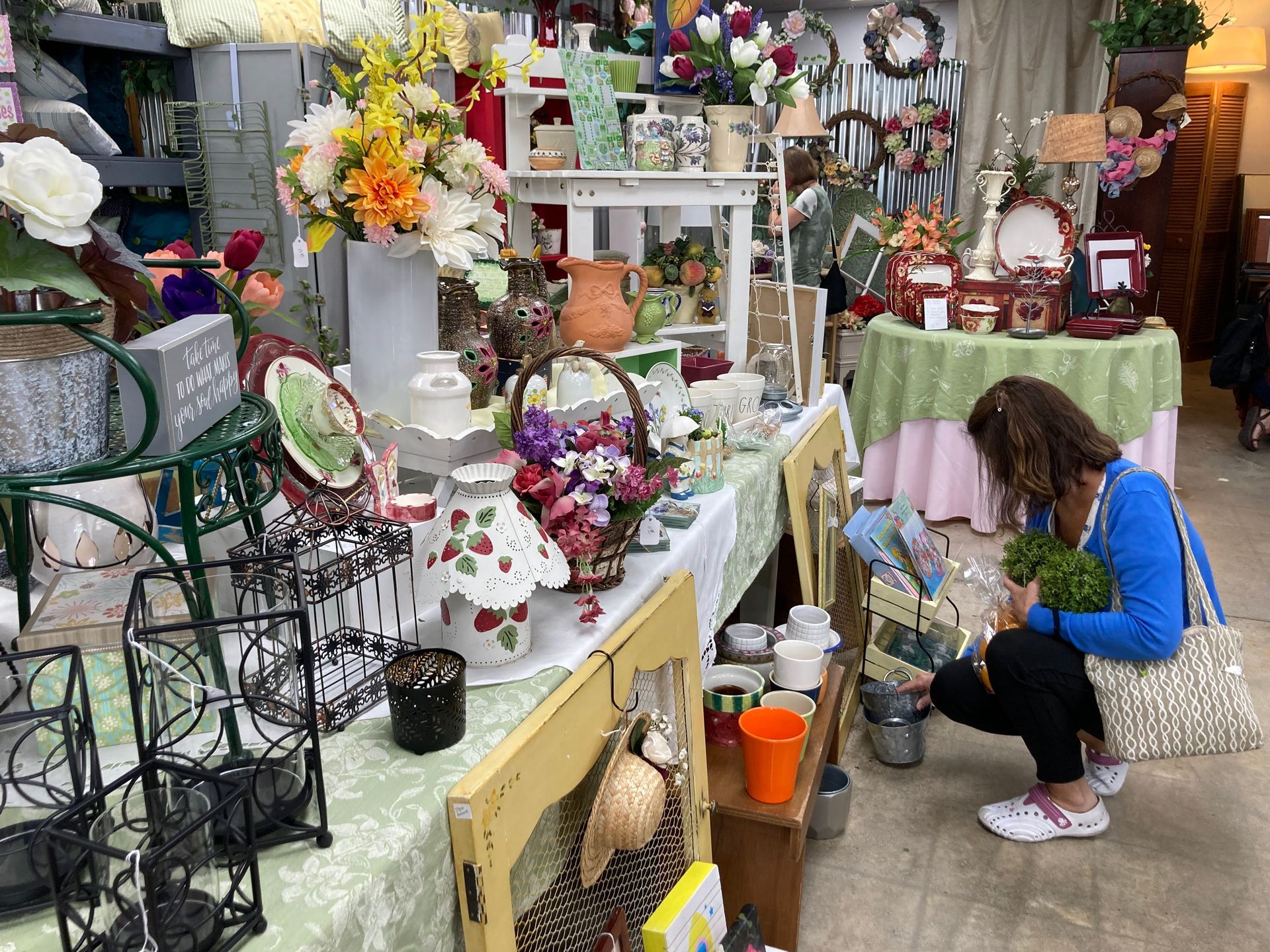 Woman shopping at a cluttered shop filled with flowers, decor, and ceramics. She examines a small green plant.