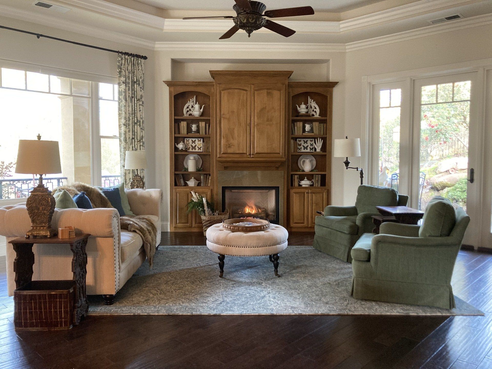 Living room with fireplace, bookshelves, green armchairs, and a beige sofa, with a rug on a dark wood floor.