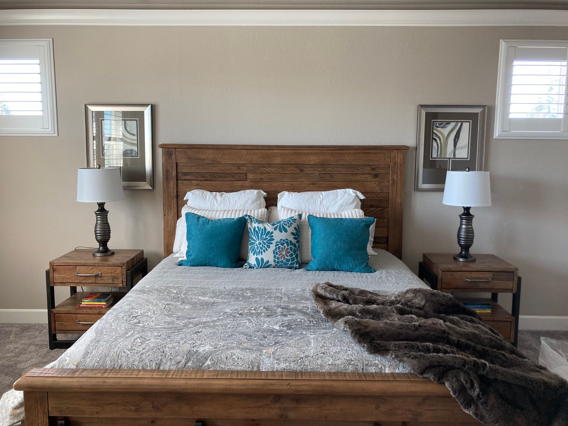 Bedroom with wooden bed, nightstands, lamps, art, and turquoise pillows against a tan wall.