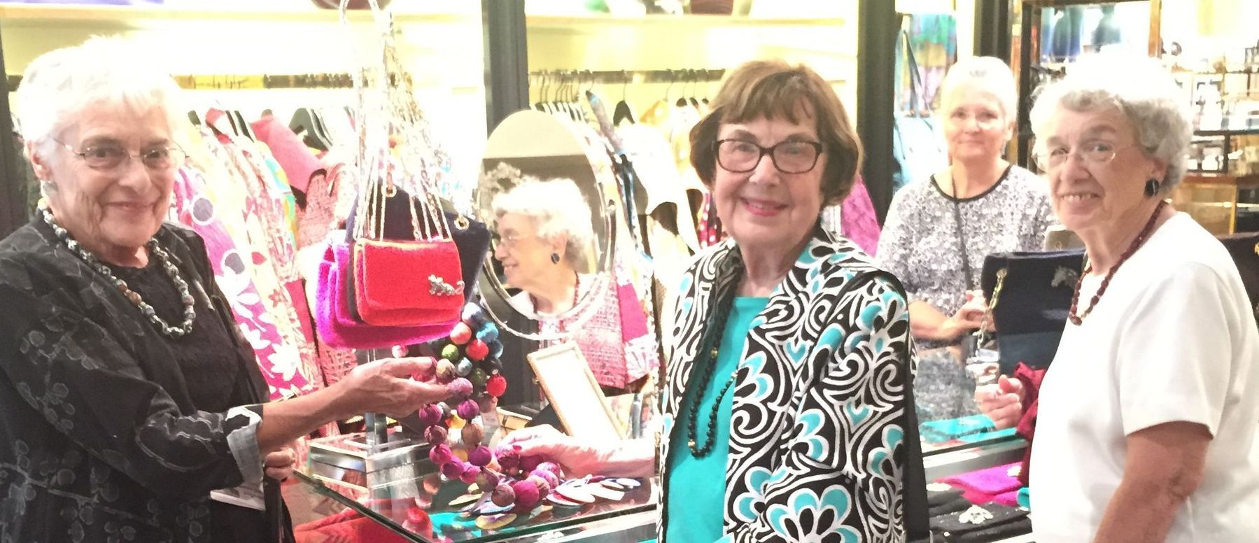 Four older women in a shop, smiling at the camera. They are surrounded by jewelry and bags.