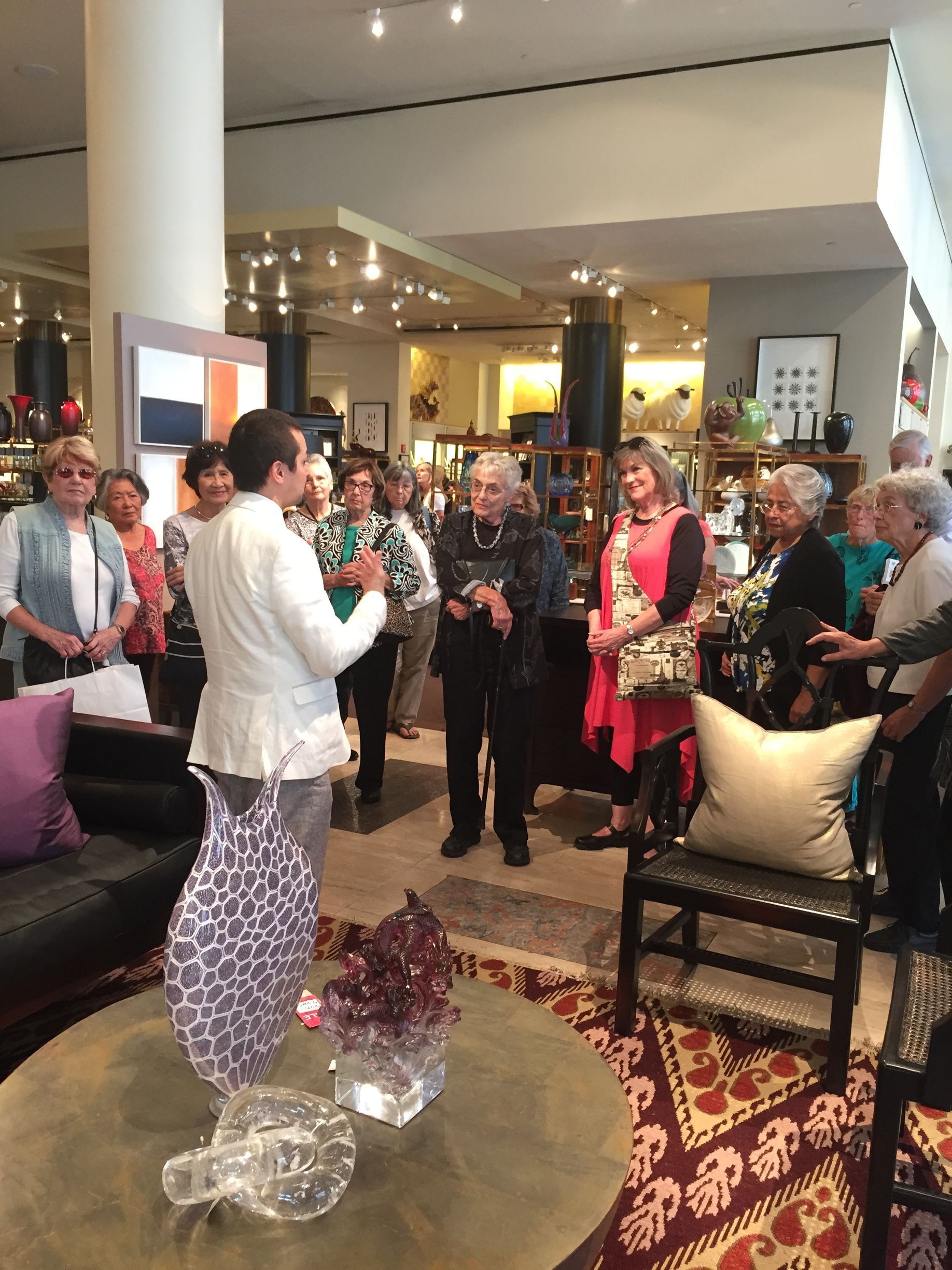 A group of women listens to a man in a furniture store, looking at art and home goods.
