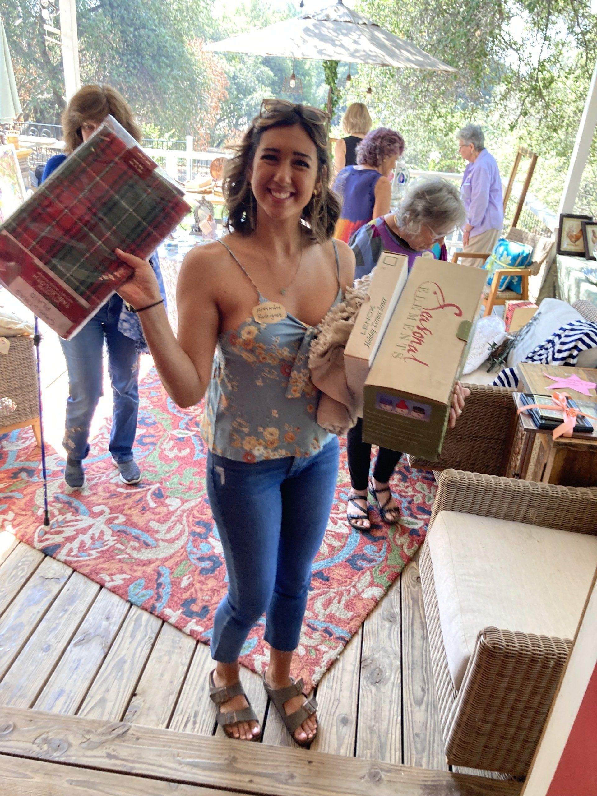 Woman holding items at an outdoor sale, smiling. Wearing floral top, jeans, sandals, with others browsing in background.