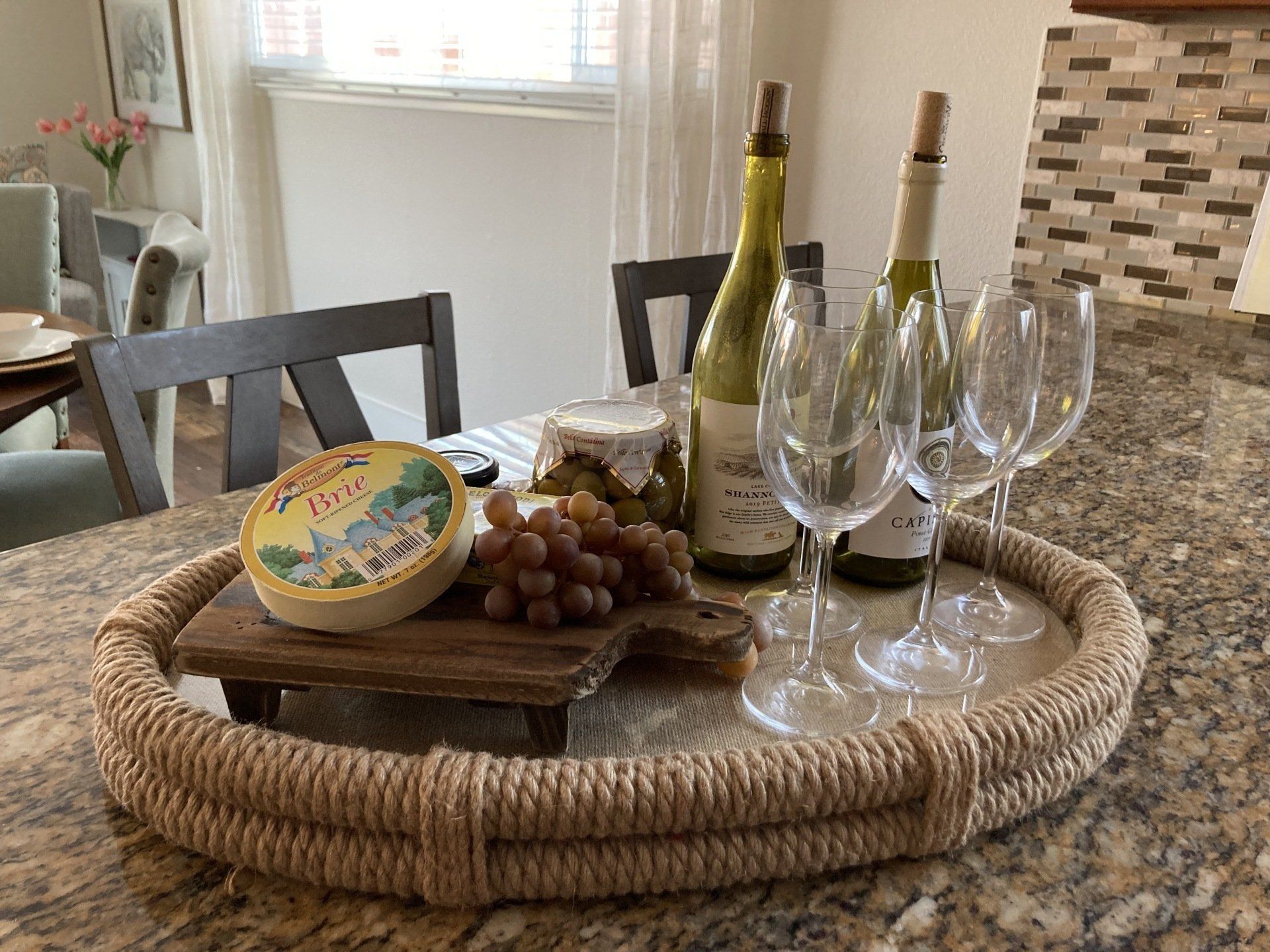 A round tray with cheese, grapes, wine bottles, glasses, and pickles on a countertop.