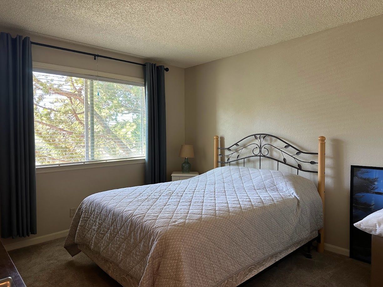 Bedroom with bed, window with curtains, and a nightstand. Beige walls and patterned bedspread.