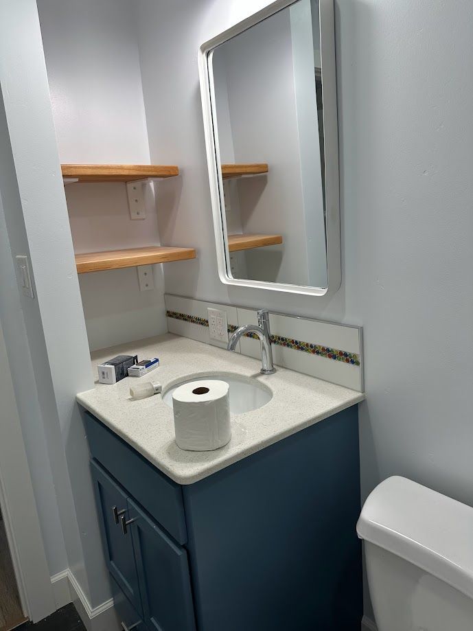 Small blue bathroom with a vanity, toilet, mirror, and shelving. White walls, toilet paper roll on counter.