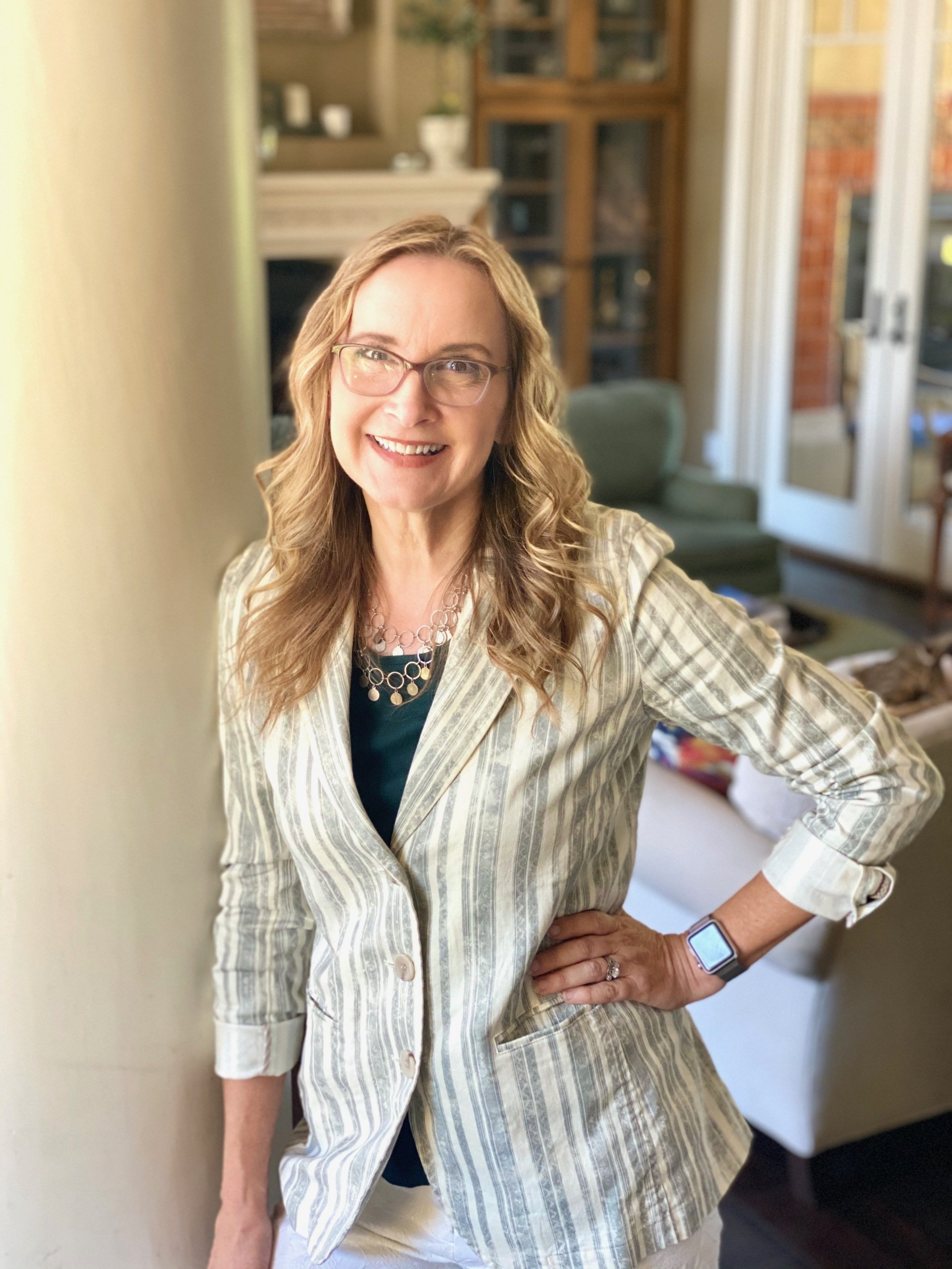 Woman with glasses in a blazer and white pants, smiling, posing by a pillar in a living room.
