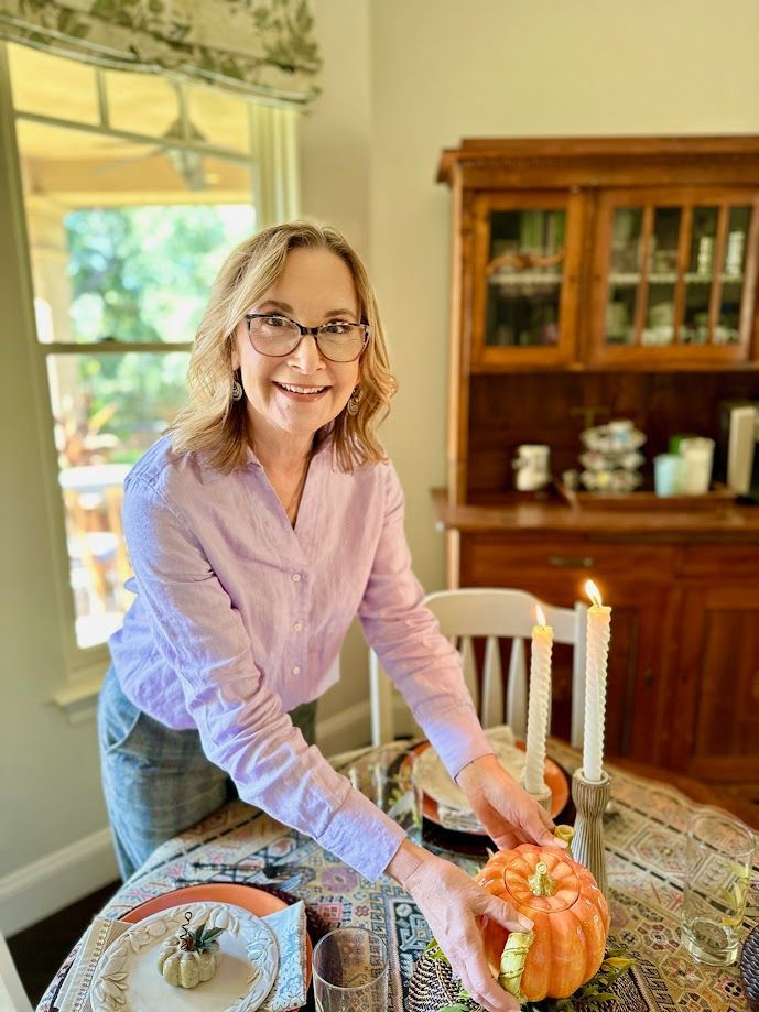 Woman arranging a pumpkin centerpiece on a dining table beside lit candles and autumn decor