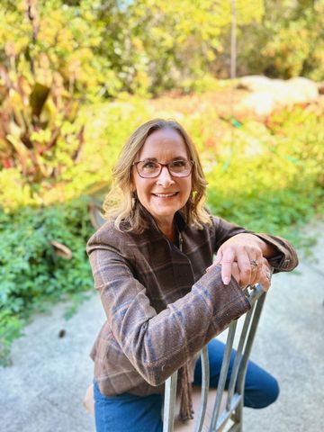 Smiling woman with glasses sitting outdoors on a chair in a sunny garden.