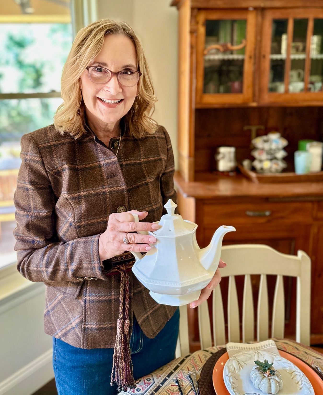 Woman in glasses, holding white teapot, smiles. Wearing plaid jacket and jeans, standing in dining room.
