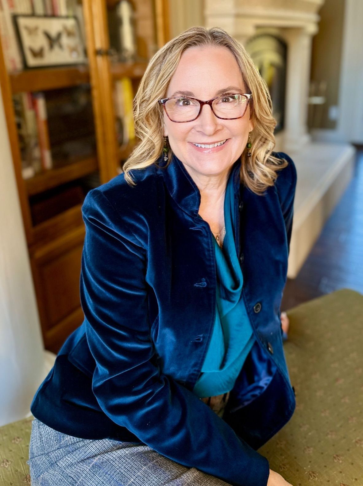Woman with glasses smiles, wearing blue velvet jacket. Leans on a cushion indoors with a fireplace in the background.