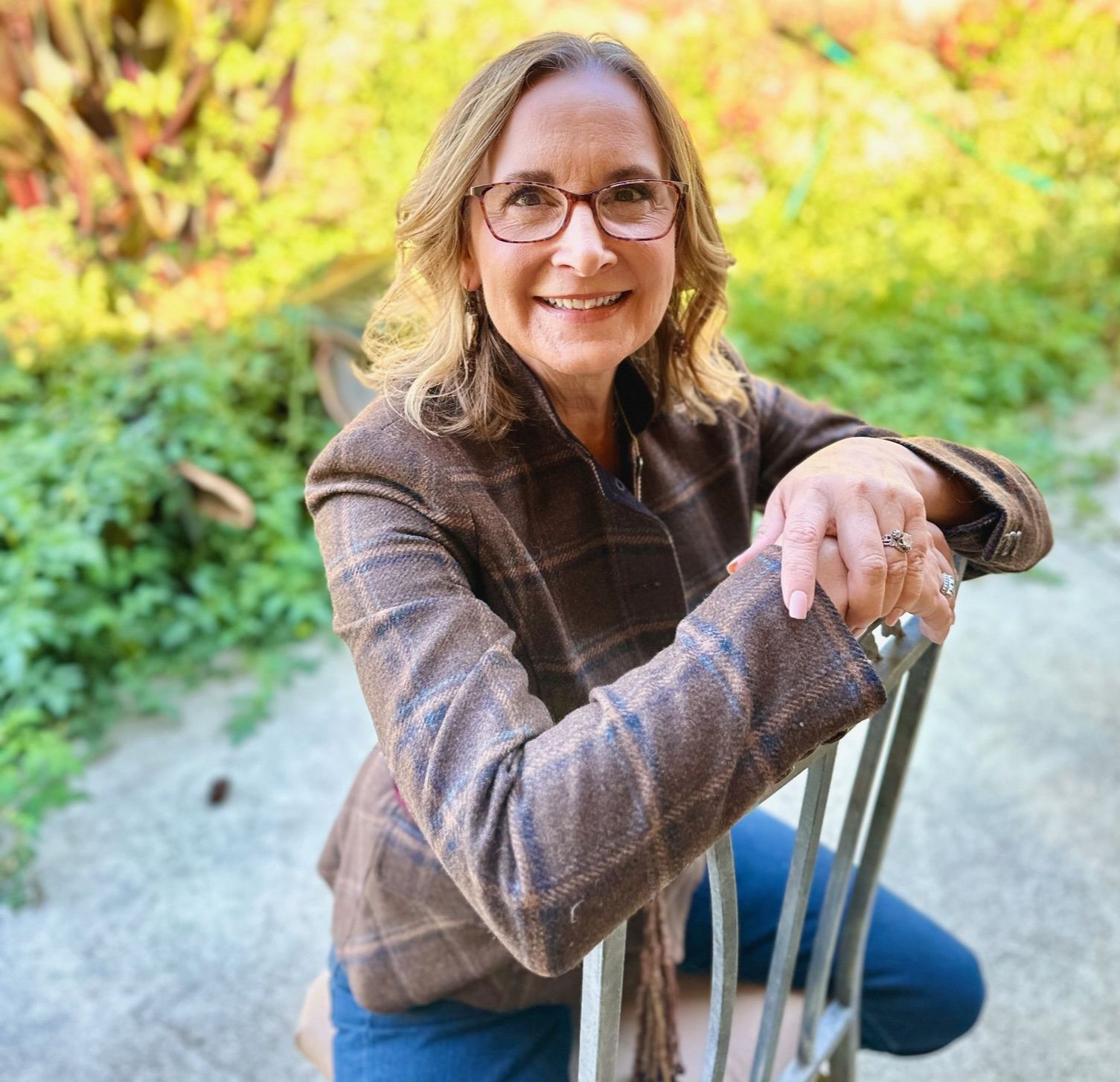 Woman with glasses in a brown blazer sits on a chair outdoors, smiling.