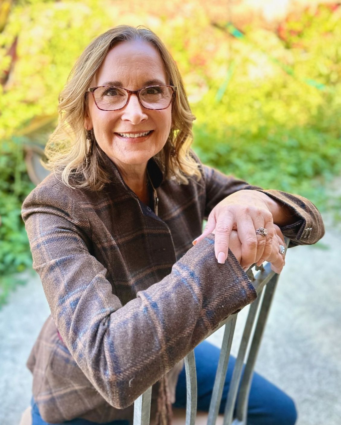 Woman in brown blazer and glasses smiles, seated outdoors.