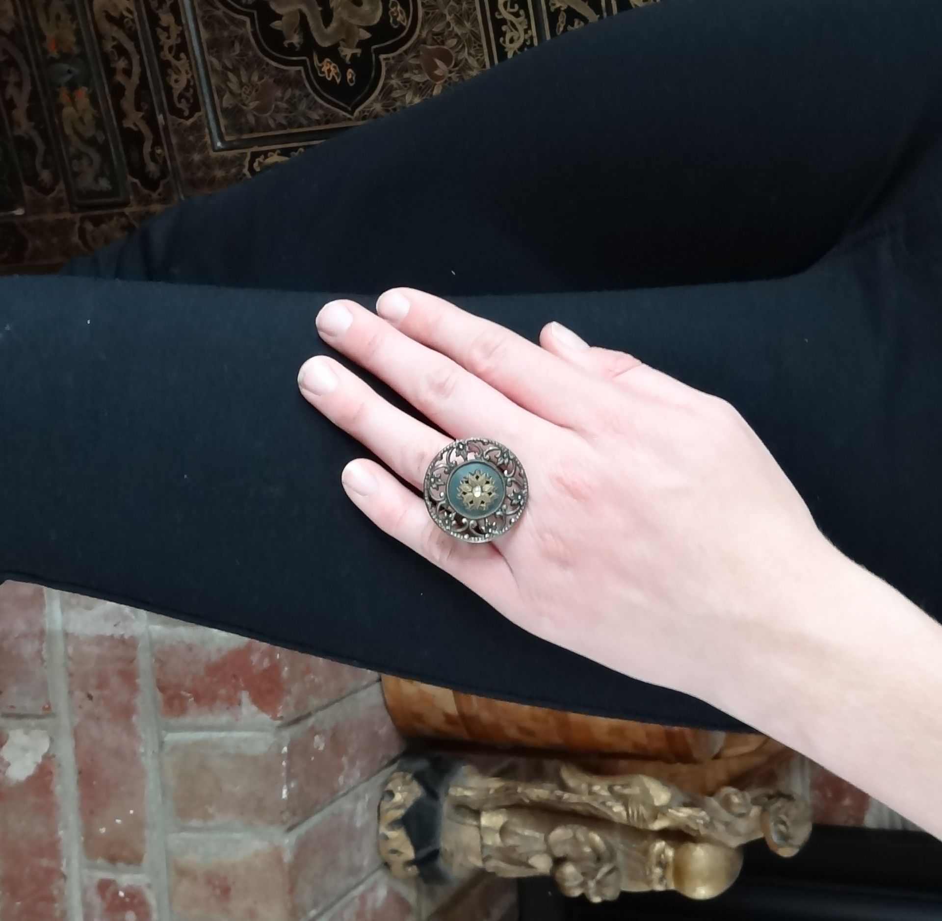 Hand wearing a large decorative ring, resting on black pants near a fireplace.