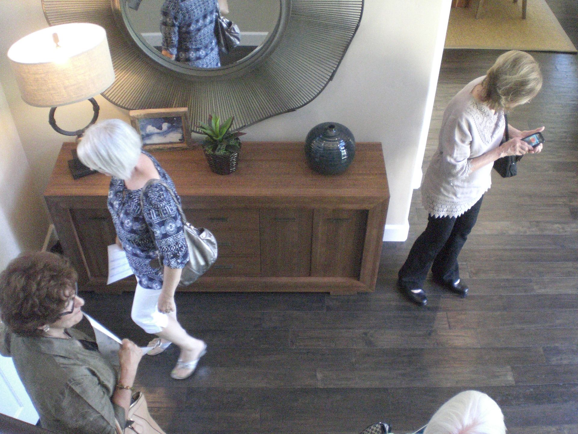 Three women in a home interior; one takes a photo, another walks, and the last sits.