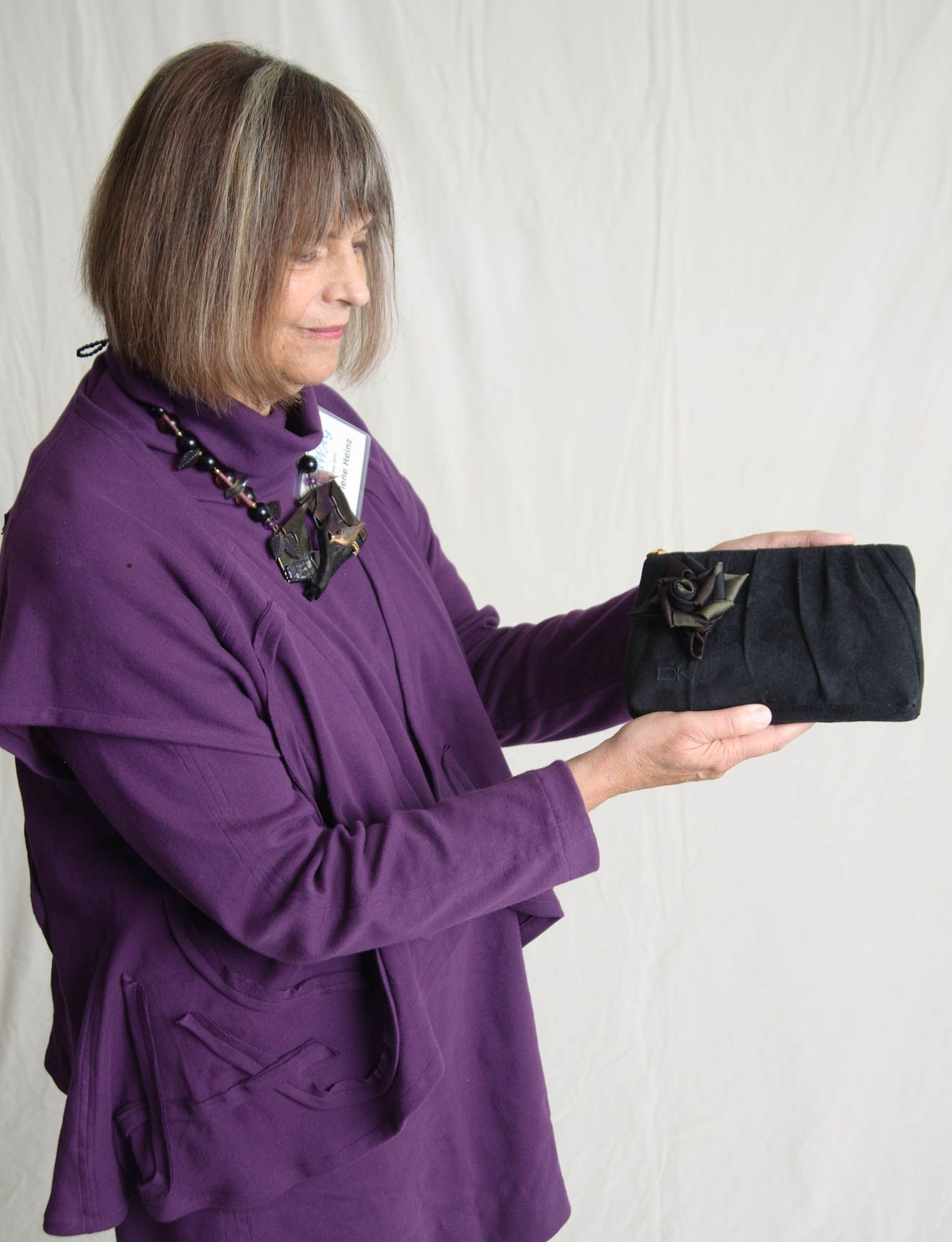 Woman in purple holds a black clutch, stands against a white backdrop.