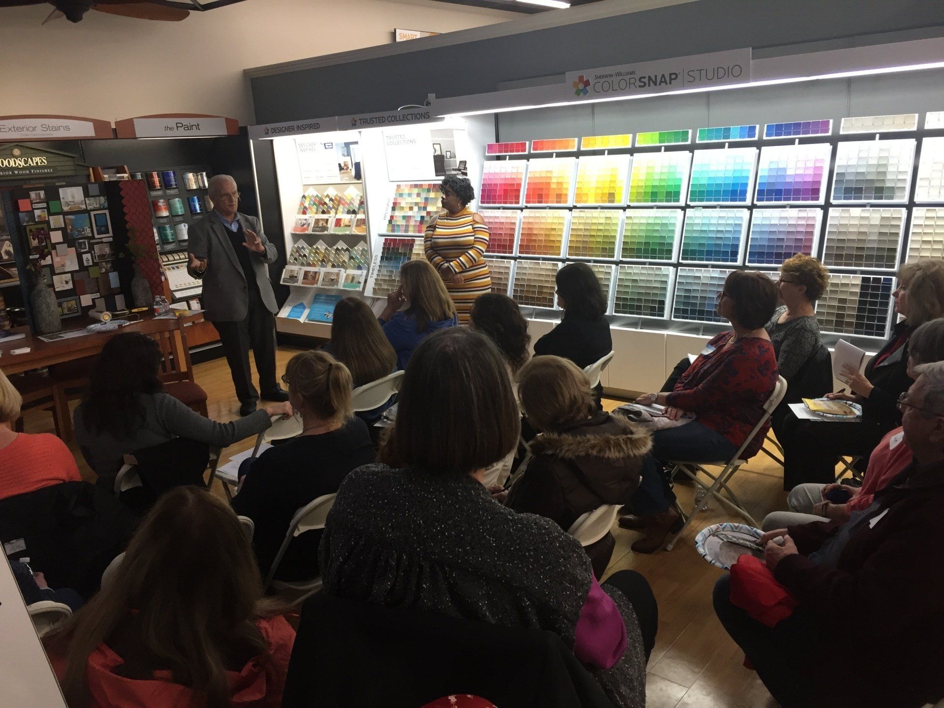 A man speaks to a seated audience in a paint store. Paint swatches are behind him.