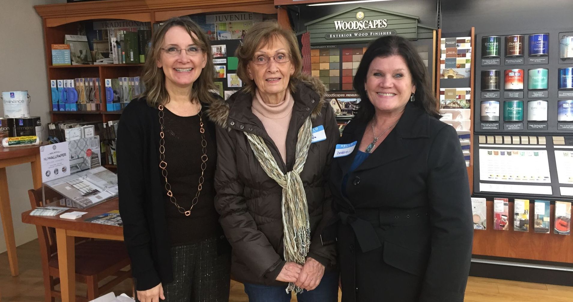 Three women smiling in a store. One in a brown coat, the others in dark outfits. Jewelry display in background.