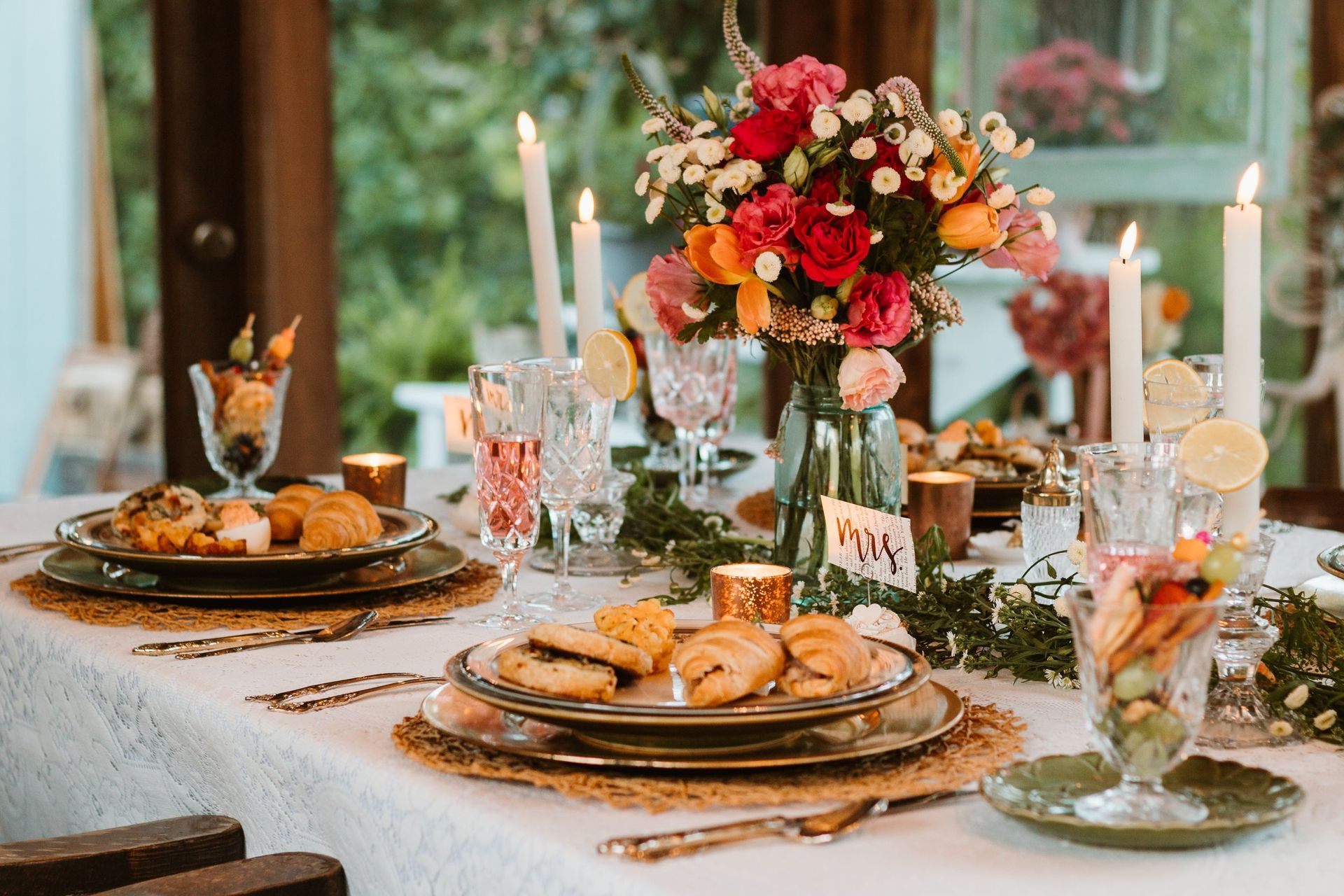 Elegant table setting with flowers, candles, food, and drinks in a bright room.