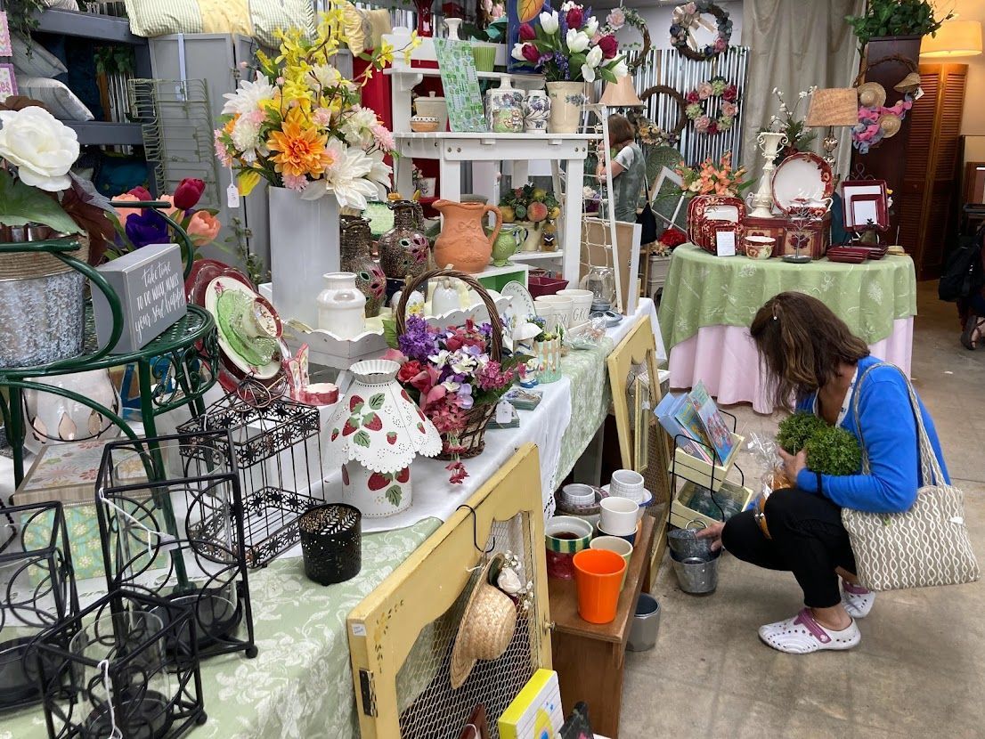 Woman browses a cluttered shop display of flowers, dishes, and decor.