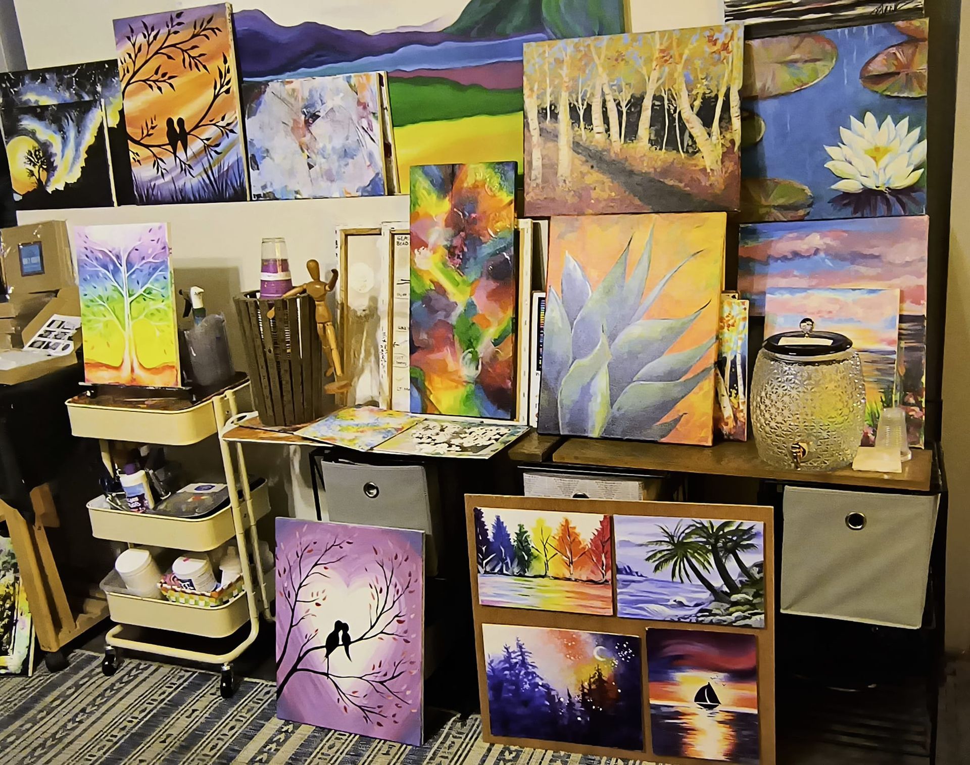 An artist's studio, walls covered in colorful paintings of landscapes, a cart with art supplies, and a storage shelf.