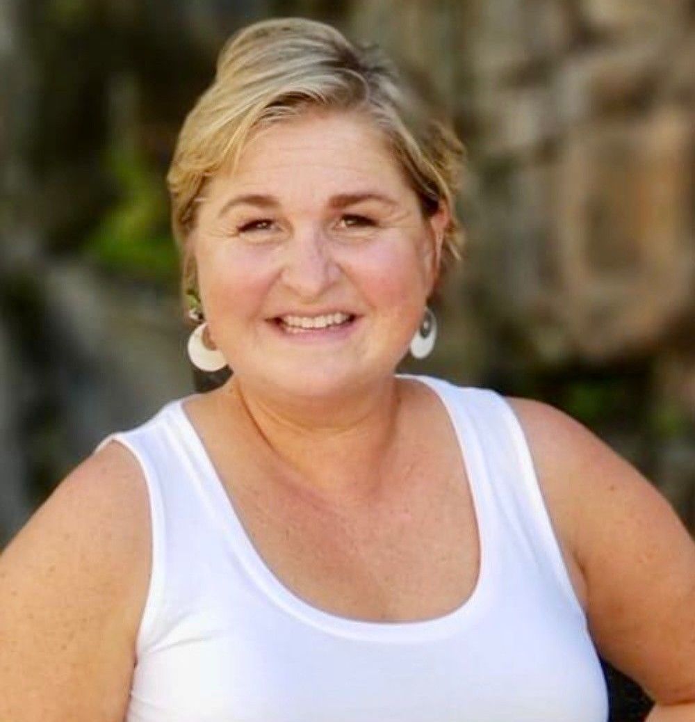 Blonde woman smiles, wearing a white tank top and earrings outdoors.